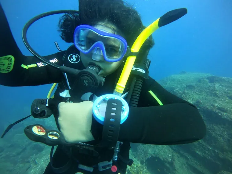 A diver underwater in scuba gear, mask and regulator on, signalling to the camera.